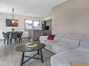A modern living room with a grey sofa, a dining table with chairs, a small coffee table, and a kitchen area with a grey countertop and cabinets.