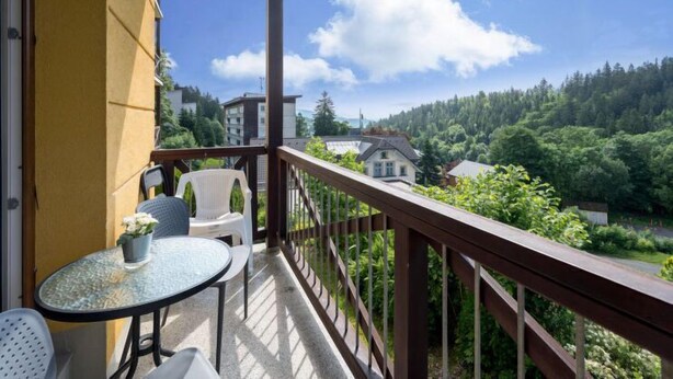 Un balcon avec une table et des chaises, donnant sur une zone boisée et des bâtiments.