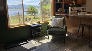 A cozy kitchen with a large window overlooking a scenic view, a green armchair, and a wood-burning stove.