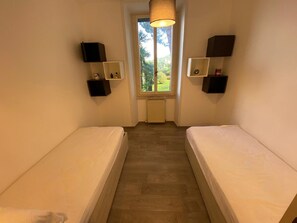 Une pièce étroite avec deux lits simples, une fenêtre donnant sur un paysage verdoyant et des haut-parleurs fixés au mur.