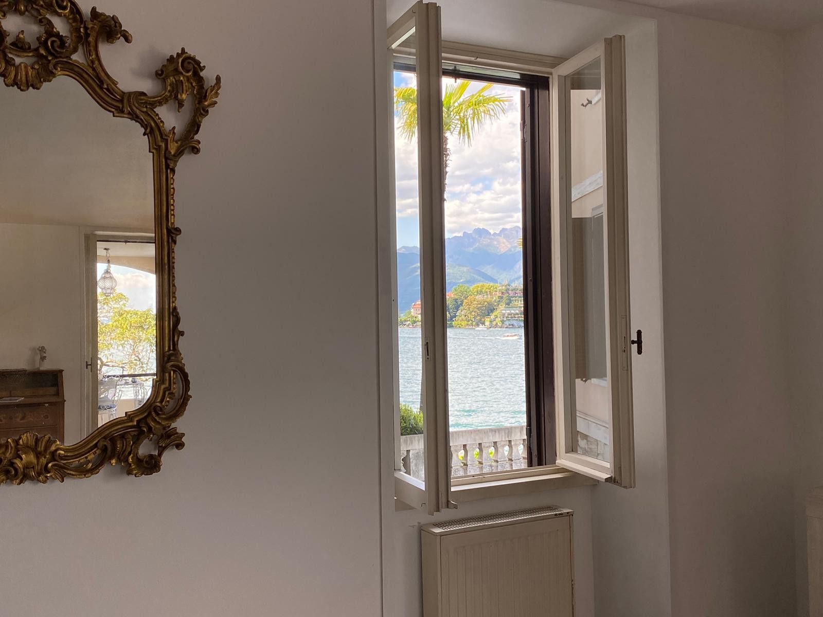 Una stanza con un grande specchio decorato, una finestra vista lago e montagne, e un termosifone.