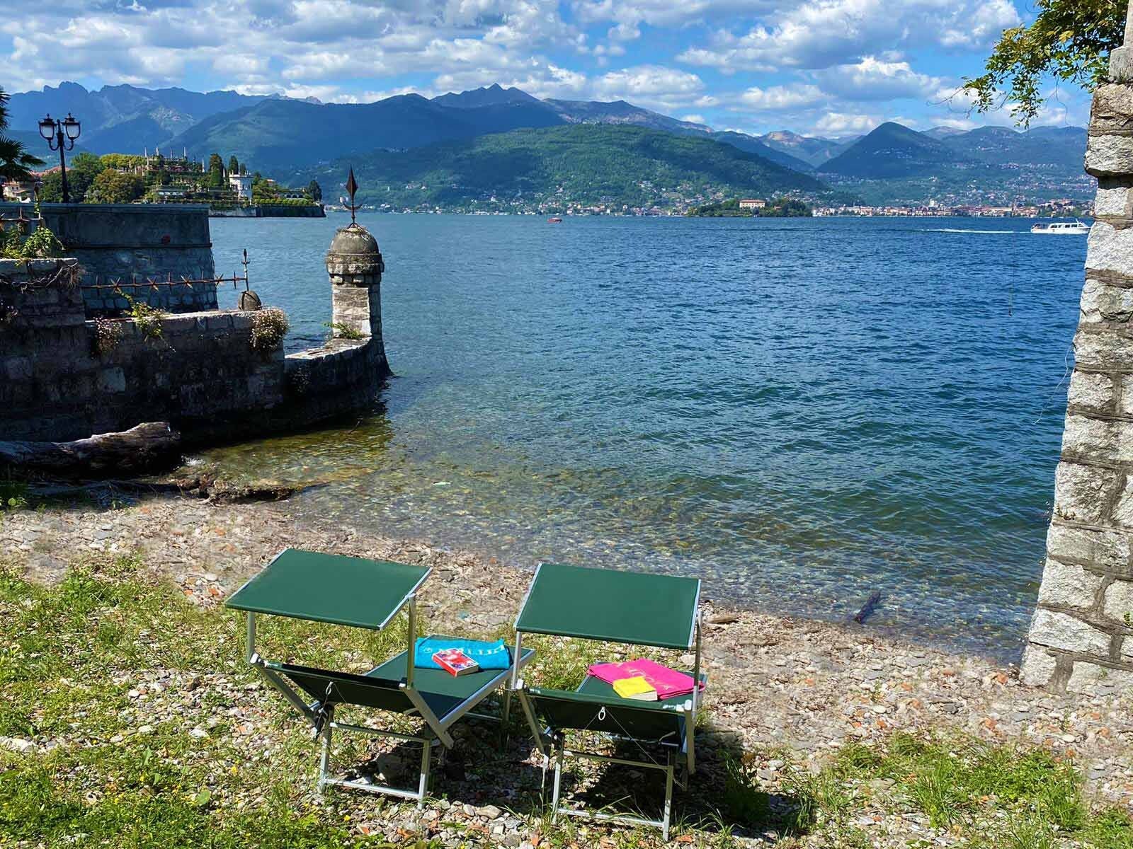 Vista sul lago con una costruzione in pietra, due tavoli verdi con libri e cielo sereno.