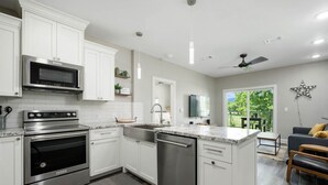 Une cuisine moderne avec des meubles blancs, des appareils en acier inoxydable, un lave-vaisselle et un coin repas comprenant un canapé gris et un banc en bois.