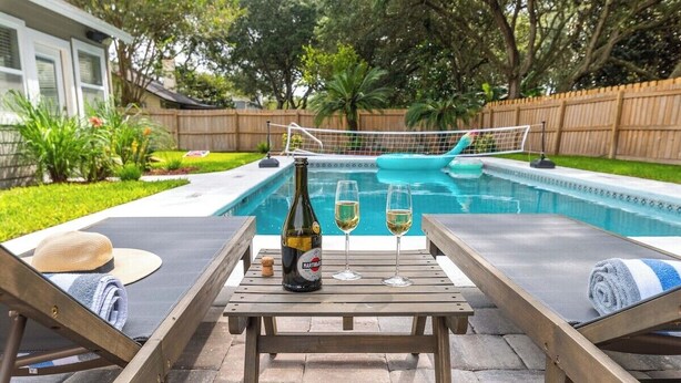 Un espace aménagé au bord de la piscine, avec des chaises longues, une table sur laquelle se trouvent une bouteille et des verres, et un filet de volley-ball.