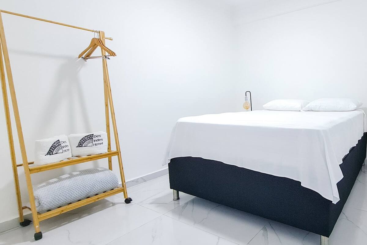 A neatly arranged hotel room with a bed, a wooden hanger rack with towels, a bedside table with a lamp, and a door.