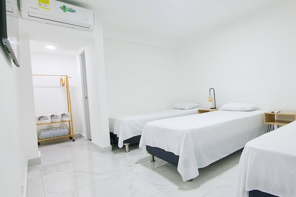 A hotel room with three single beds, a wooden nightstand, a small table, and a shelf with boxes.