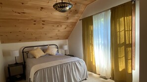 Un dormitorio con techo de madera, una cama con colcha a rayas, una lámpara de noche y una ventana con cortinas translúcidas.