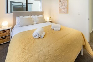 Una cama bien hecha con una colcha color beige, dos almohadas y toallas enrolladas sobre una mesita de noche.