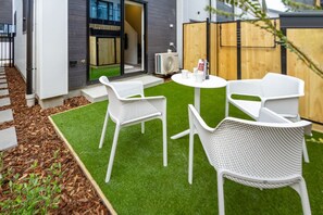 Un patio con sillas de ratán blanco y una mesa, rodeado de césped artificial y una valla de madera.