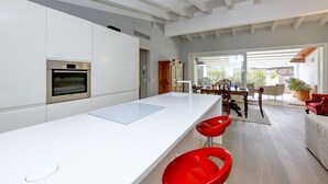 Una cocina moderna con armarios blancos, horno empotrado y una isla grande con encimera blanca. Hay taburetes rojos y un comedor con mesa y sillas.