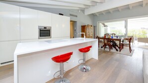 Una cocina moderna con armarios blancos, una isla central con taburetes rojos y una zona de comedor con sillas de madera.
