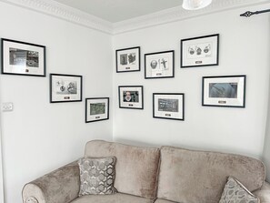 Een woonkamer met een beige bank, een kussen met een patroon en een muur versierd met zwart-wit foto's in lijsten.