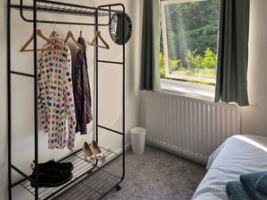 Et værelse med en seng, et vindue med udsigt til grønne omgivelser, en radiator og et tøjstativ med sko og bøjler.