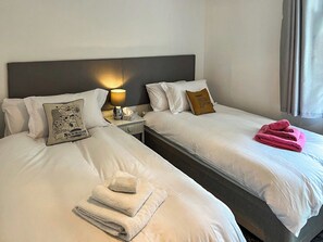 Deux lits avec du linge de lit blanc, une table de chevet avec une lampe, un oreiller orné d’une carte et une serviette pliée.