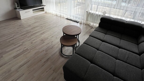 A modern living room with a large sectional sofa, a flat-screen TV on a white stand, two round wooden tables, and sheer curtains over a large window.