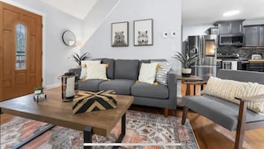 Un salon moderne avec un canapé gris, une table basse en bois et un tapis à motifs. On aperçoit également un coin cuisine avec des appareils en acier inoxydable.