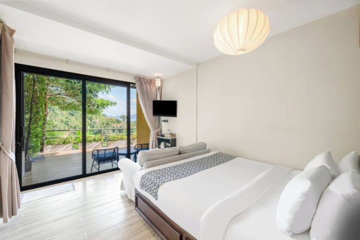 A bedroom with a large bed, a TV, and a balcony with a view of greenery.