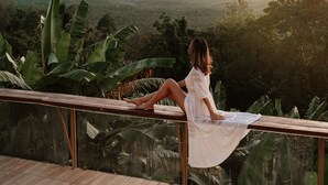 Een vrouw zittend op een balkon met uitzicht op een bergachtig landschap, een grote rotsformatie en een wateroppervlak.