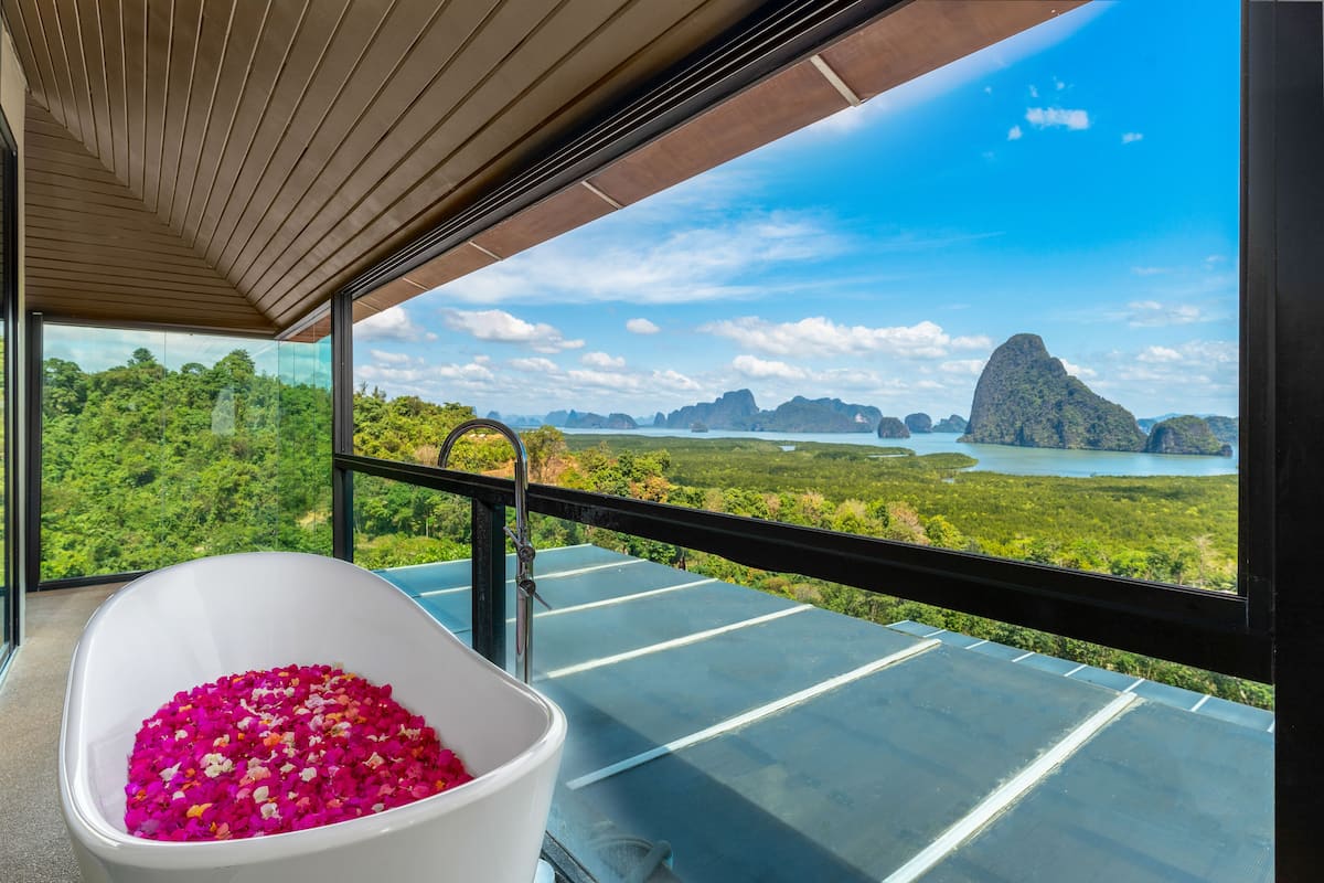 Een moderne badkamer met een groot bad vol roze bloemen, een douchecabine van glas en uitzicht op een tropisch landschap met bergen en water.