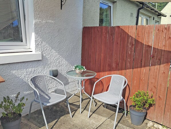 A patio with two white metal chairs and a small round table, a potted plant, and a wooden fence. - 1 bedroom accommodation in Luncarty, near Perth (Luncarty, near Perth)