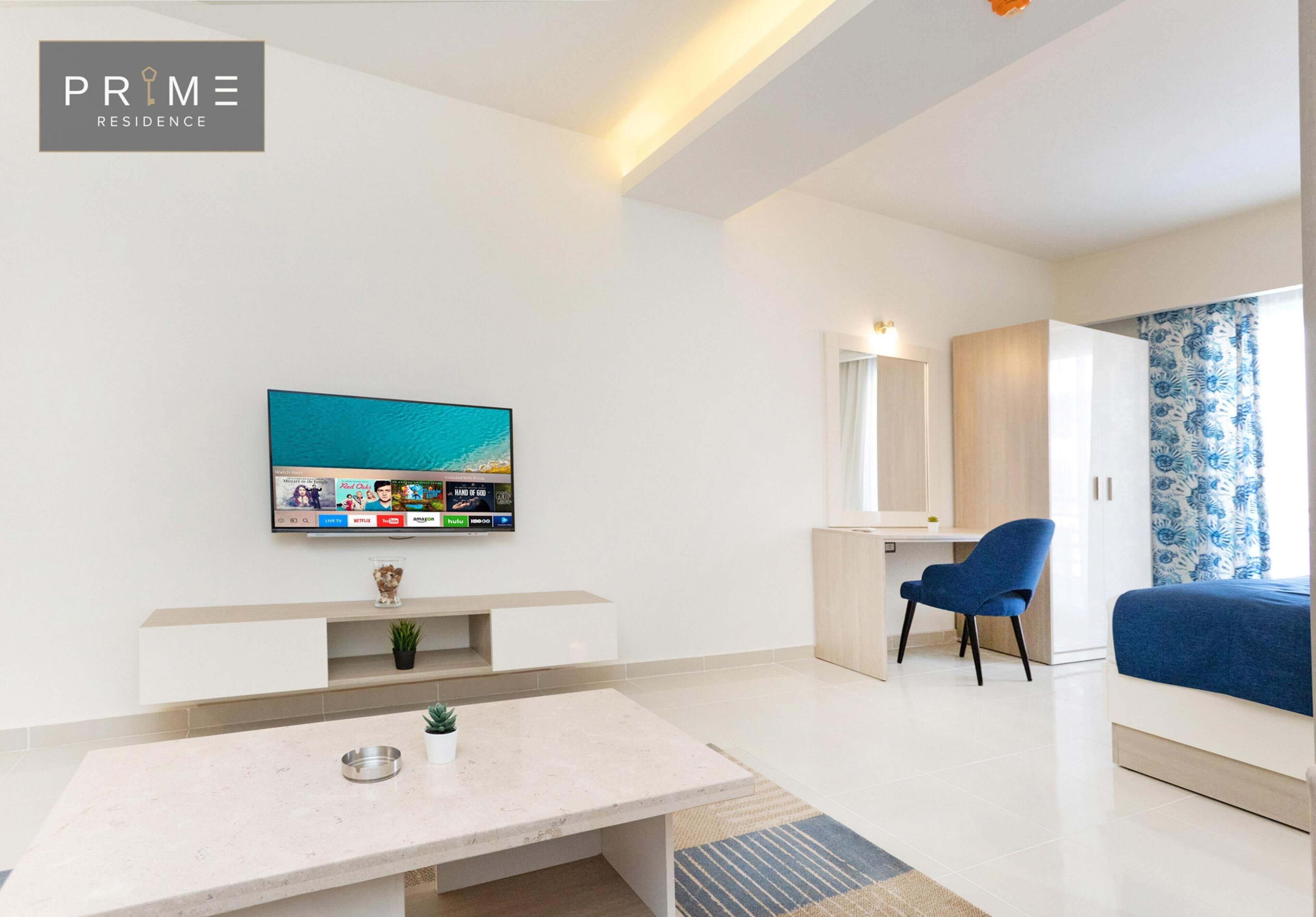 Una sala de estar moderna con una televisión de pantalla plana, una mesa de centro, un sillón azul y una cama con cortina estampada.