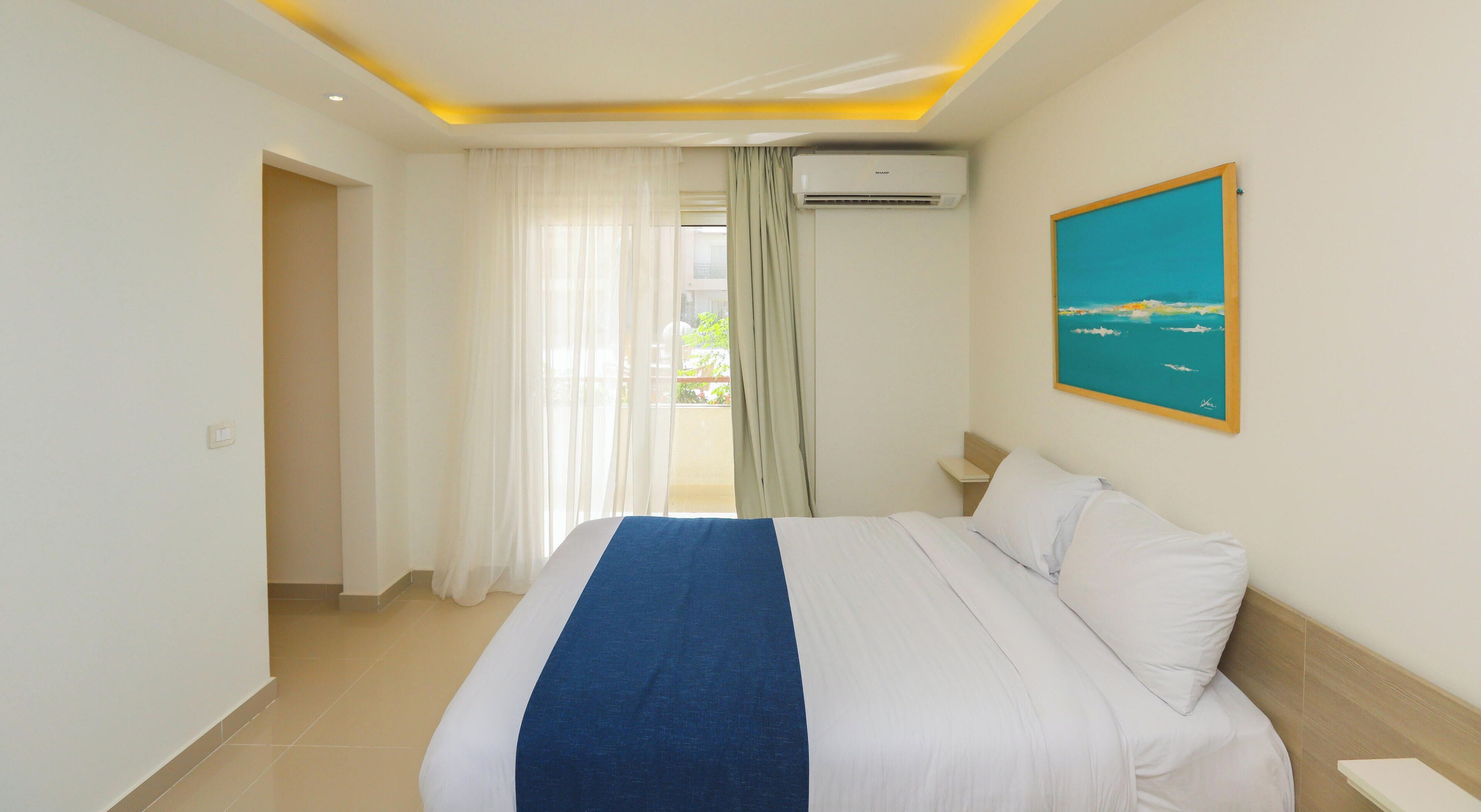 A hotel room with a bed, a painting, an air conditioning unit, and a view of the outdoors.