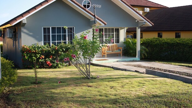 Une maison d’habitation avec un porche à l’avant, un petit jardin fleuri et une pelouse.