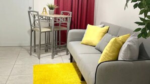 A living room with a grey sofa, yellow and grey cushions, a small table with chairs, a potted plant, and a yellow rug.