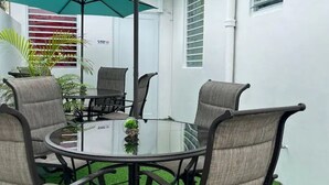 A round glass table with four chairs, surrounded by white walls and a green umbrella.