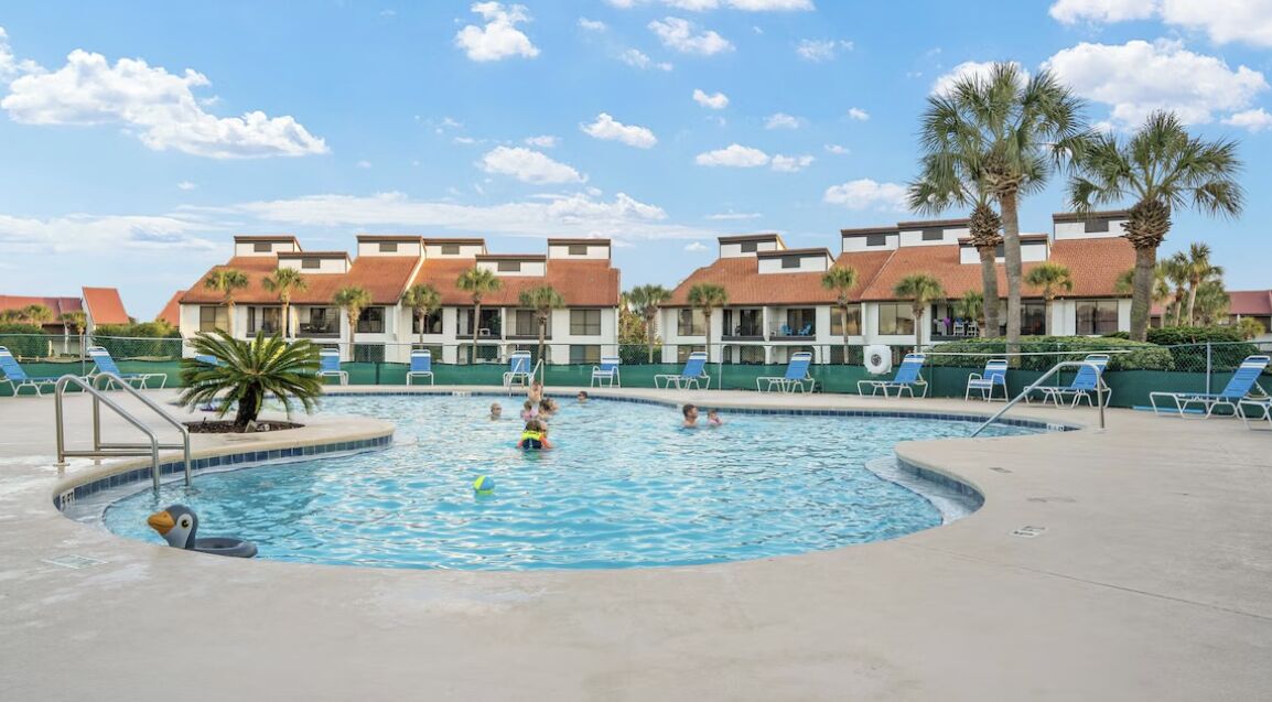 Una piscina con sillones de playa y palmeras, rodeada de edificios con tejados rojos.