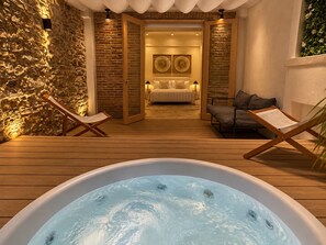 A hot tub in the foreground with a wooden deck, a stone wall on the left, and a bedroom with a bed and two fans in the background. - Unique Apartment with Jacuzzi (Ciudad de Panamá)