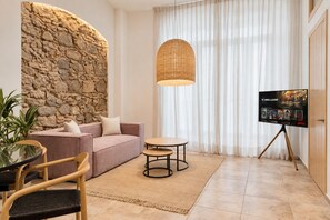 A living room with a stone wall, a sofa, a coffee table, a TV on a tripod, and a large window with sheer curtains.