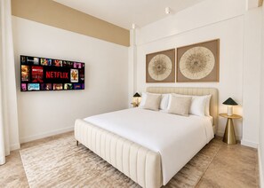 A hotel room with a large bed, two bedside tables with lamps, a wall-mounted TV displaying various shows, and two decorative wall art pieces.