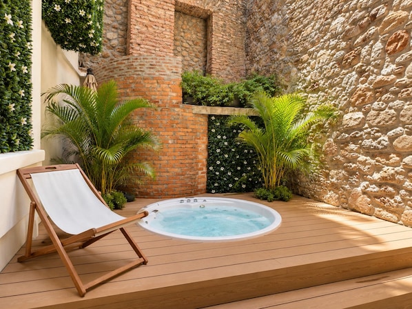 Una terraza de madera con un jacuzzi, rodeada de plantas y un muro de piedra.