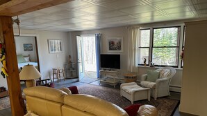 A cozy living room with a sofa, armchair, and a television. A large window provides natural light and a view of greenery outside. - The Cozy Pine, where your Grand Marais Retreat Begins! (Grand Marais)