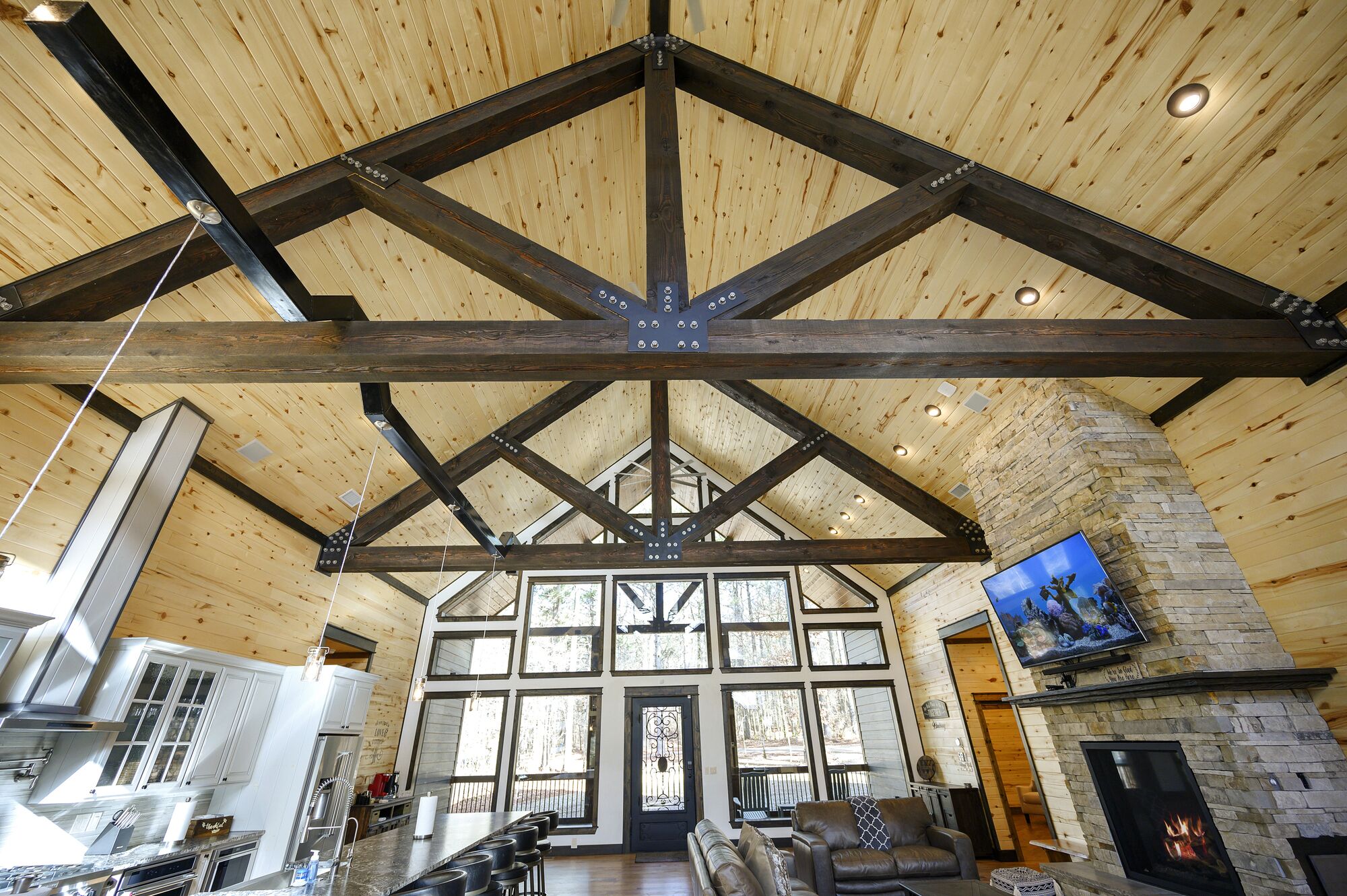A spacious living area with a high ceiling supported by wooden beams, a stone fireplace, and a large window overlooking a forest.