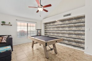 Una mesa de billar en una habitación con ventilador de techo, ventana con persianas y pared con diseño de tablones de madera.