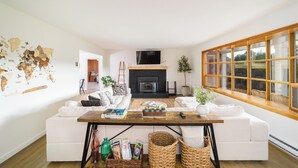 A modern living room with a fireplace, white sofas, a wooden coffee table, and a large window with a view.