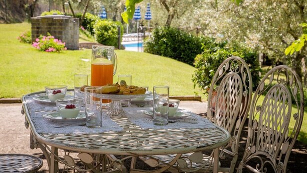 Une table à manger en plein air, avec une carafe de jus d’orange, des verres et une assiette de nourriture, entourée de chaises en osier.