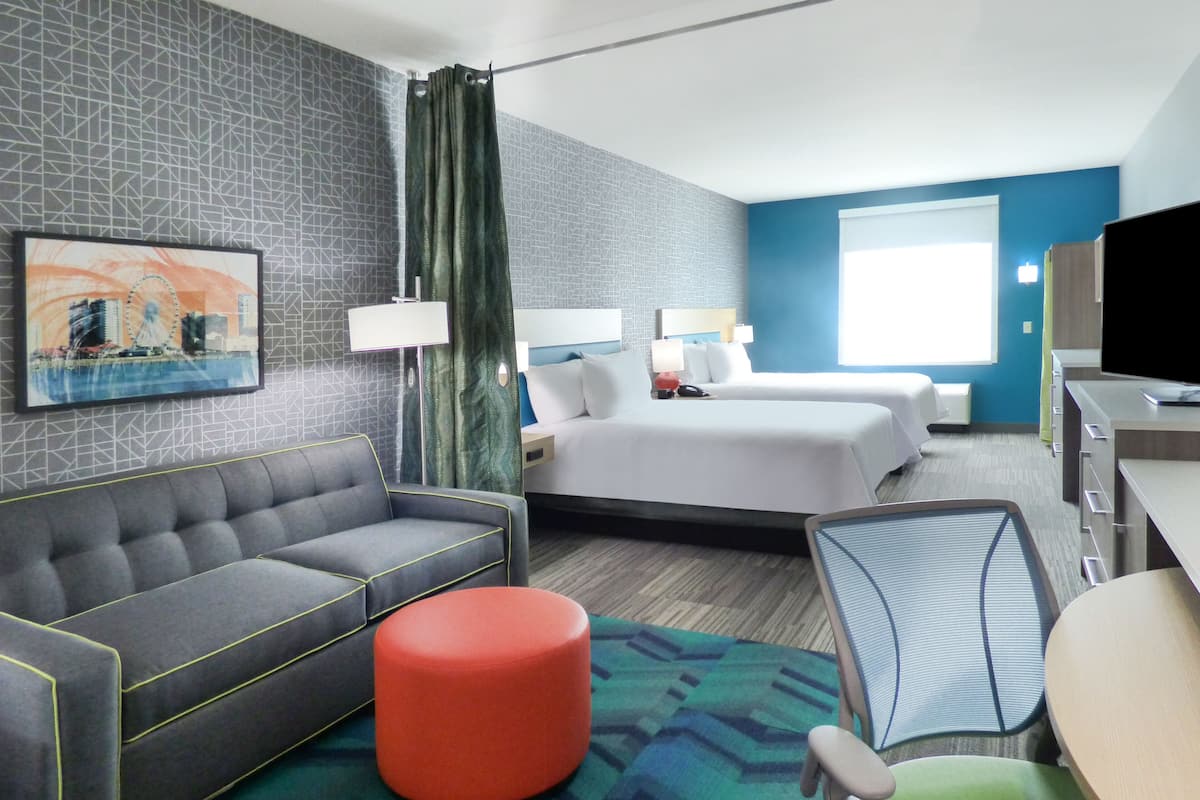 A hotel room with a grey sofa, a red ottoman, a green patterned rug, a bed with white linens, a desk with a TV, and a window with curtains.