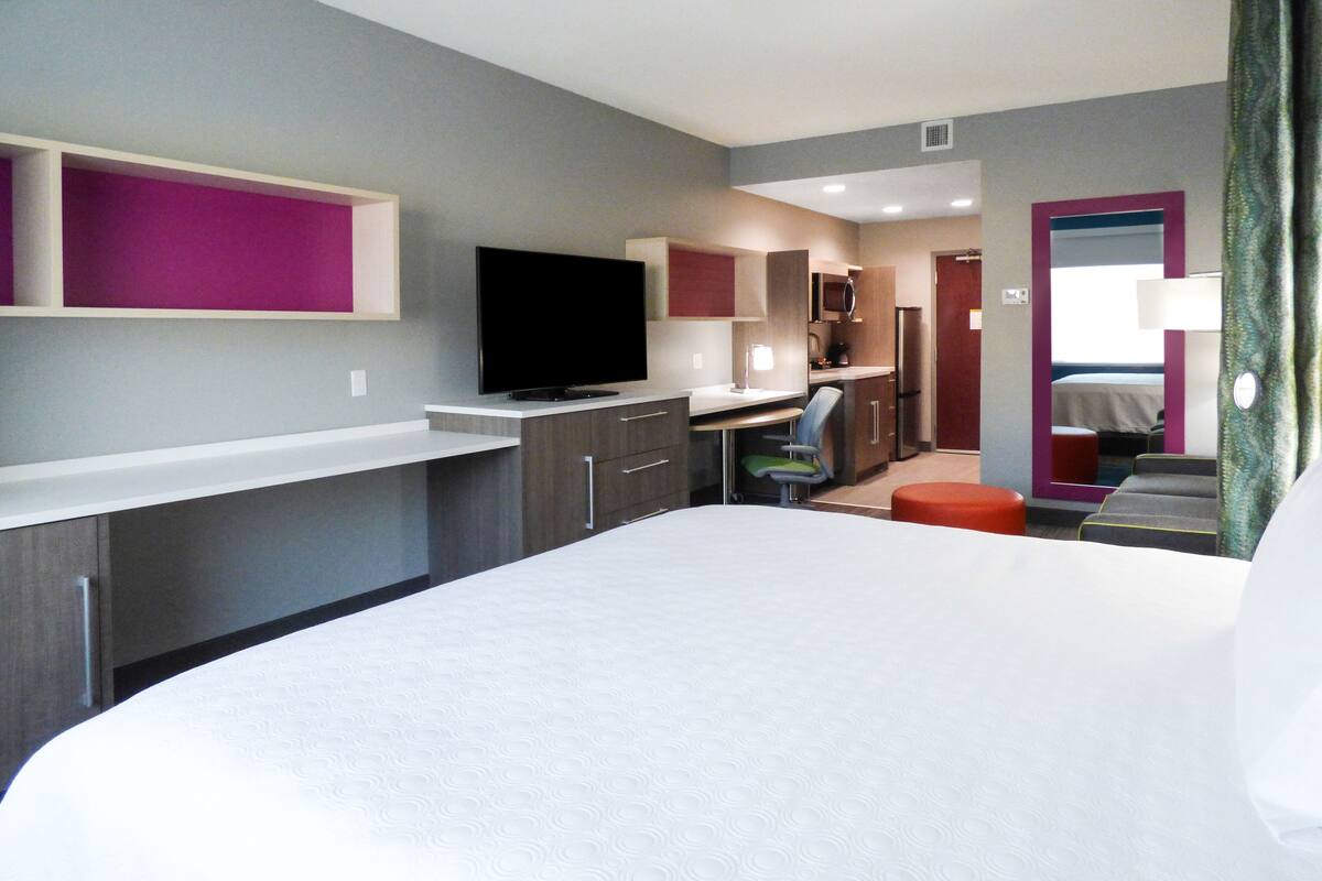 A modern hotel room with a large bed, a desk, a television, and a kitchenette.