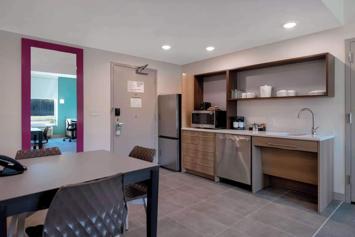 A modern kitchen with a refrigerator, microwave, and coffee maker. There is a dining table with chairs and a door leading to another room.