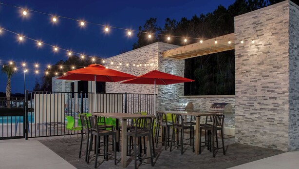 Un patio exterior con mesas y sillas, luces colgantes y una piscina al fondo.