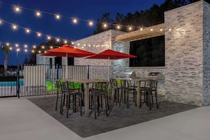 Un patio exterior con mesas y sillas, luces colgantes y una piscina al fondo.