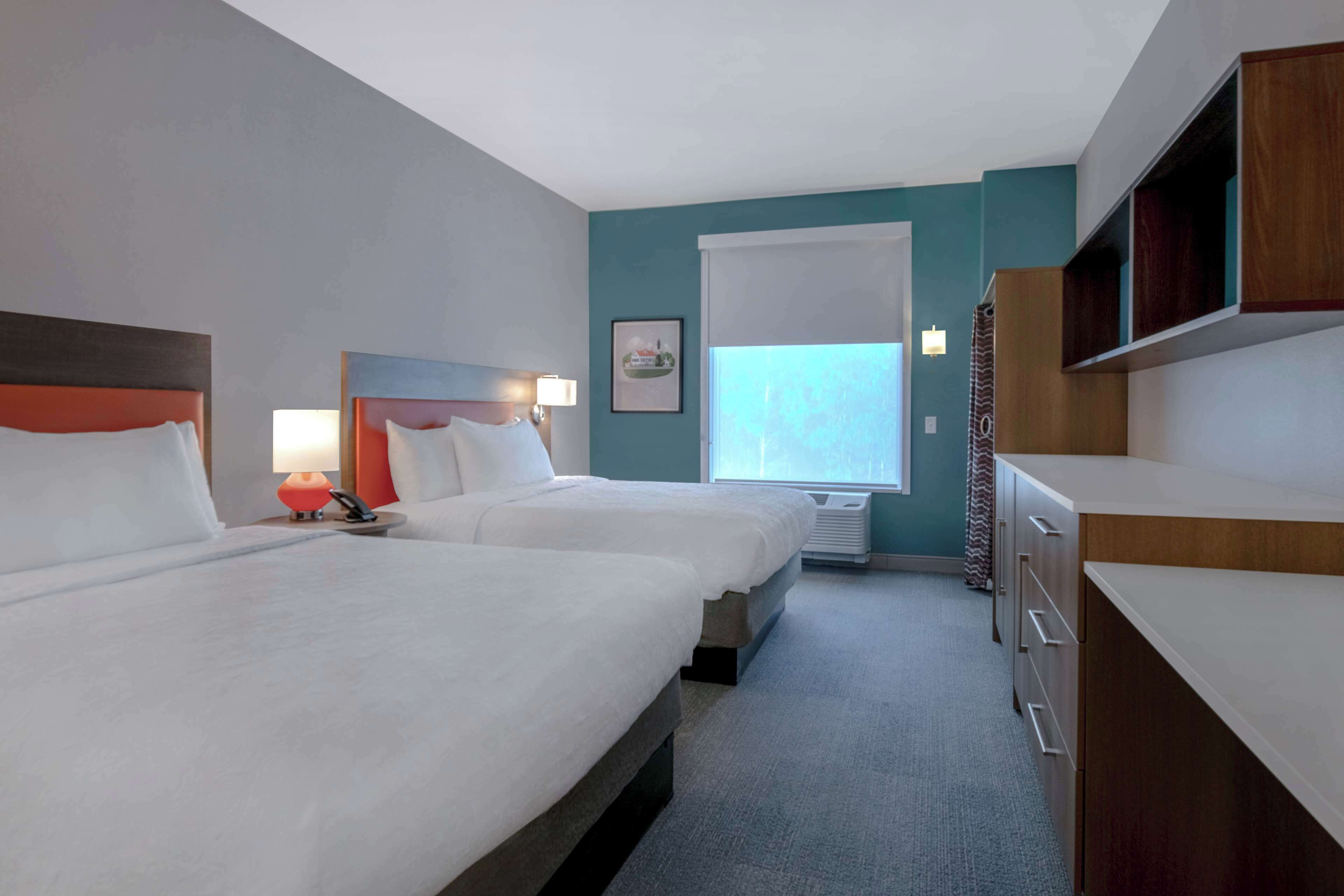 Ein Hotelzimmer mit zwei Betten, einem Schreibtisch, einem Stuhl, einer Lampe, einem Fenster und einer eingebauten Ablage.