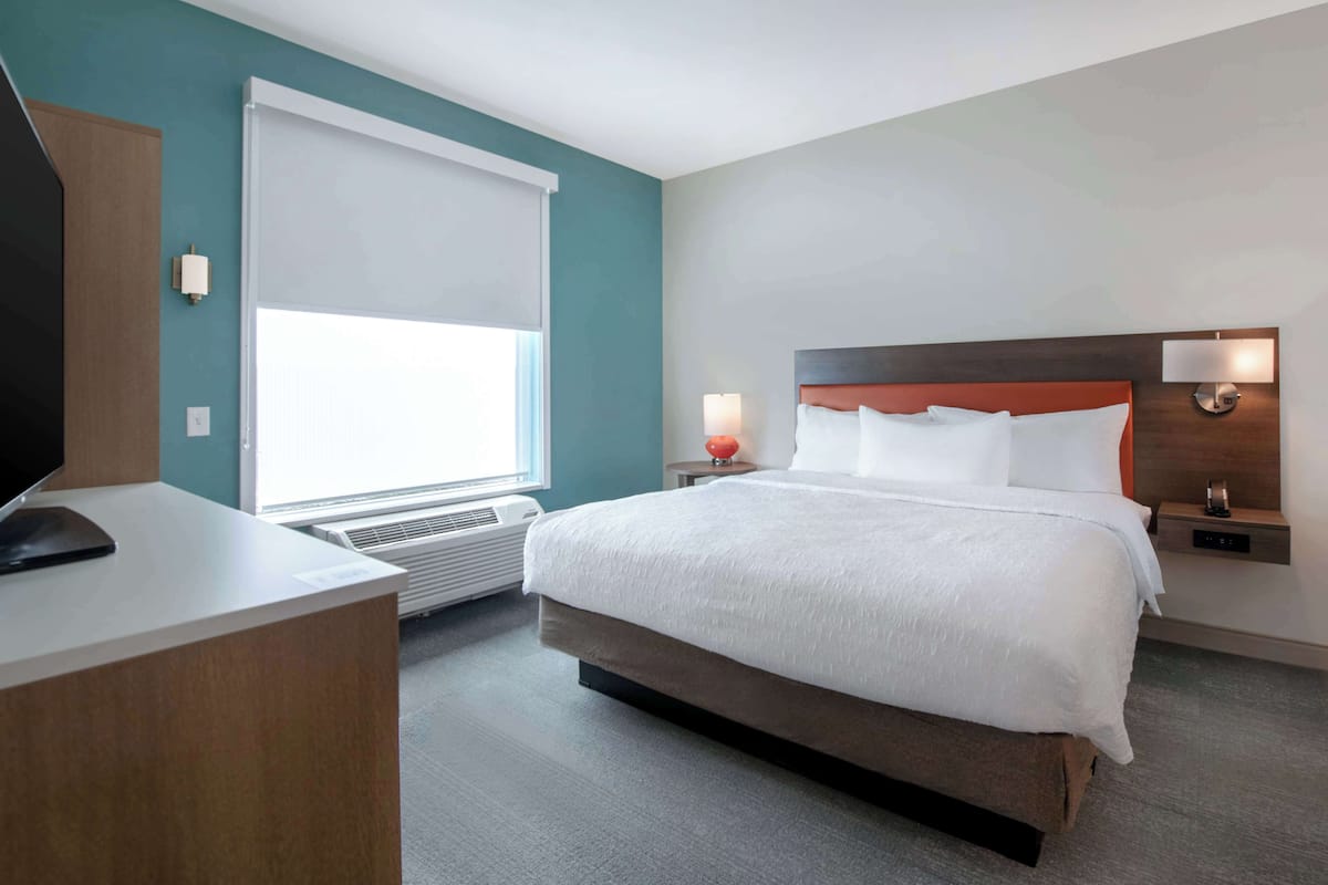 A hotel room with a large bed, a desk with a computer, a window with blinds, and a wall-mounted lamp.