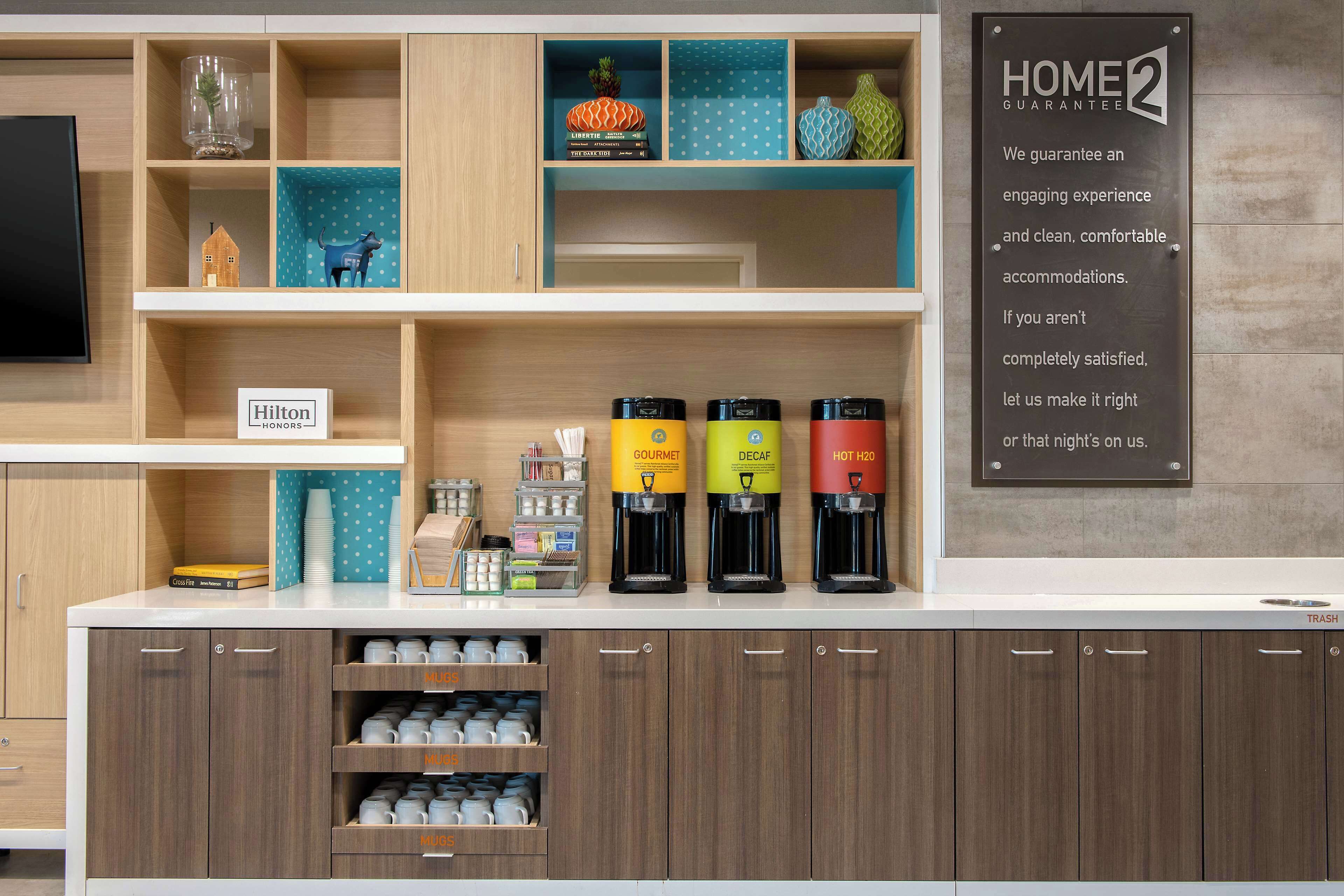 A hotel kitchenette with a 'Home 2 Guarantee' sign, a 'Hilton Honors' sign, and coffee makers labeled 'Gourmet,' 'Decaf,' and 'Hot H20.'