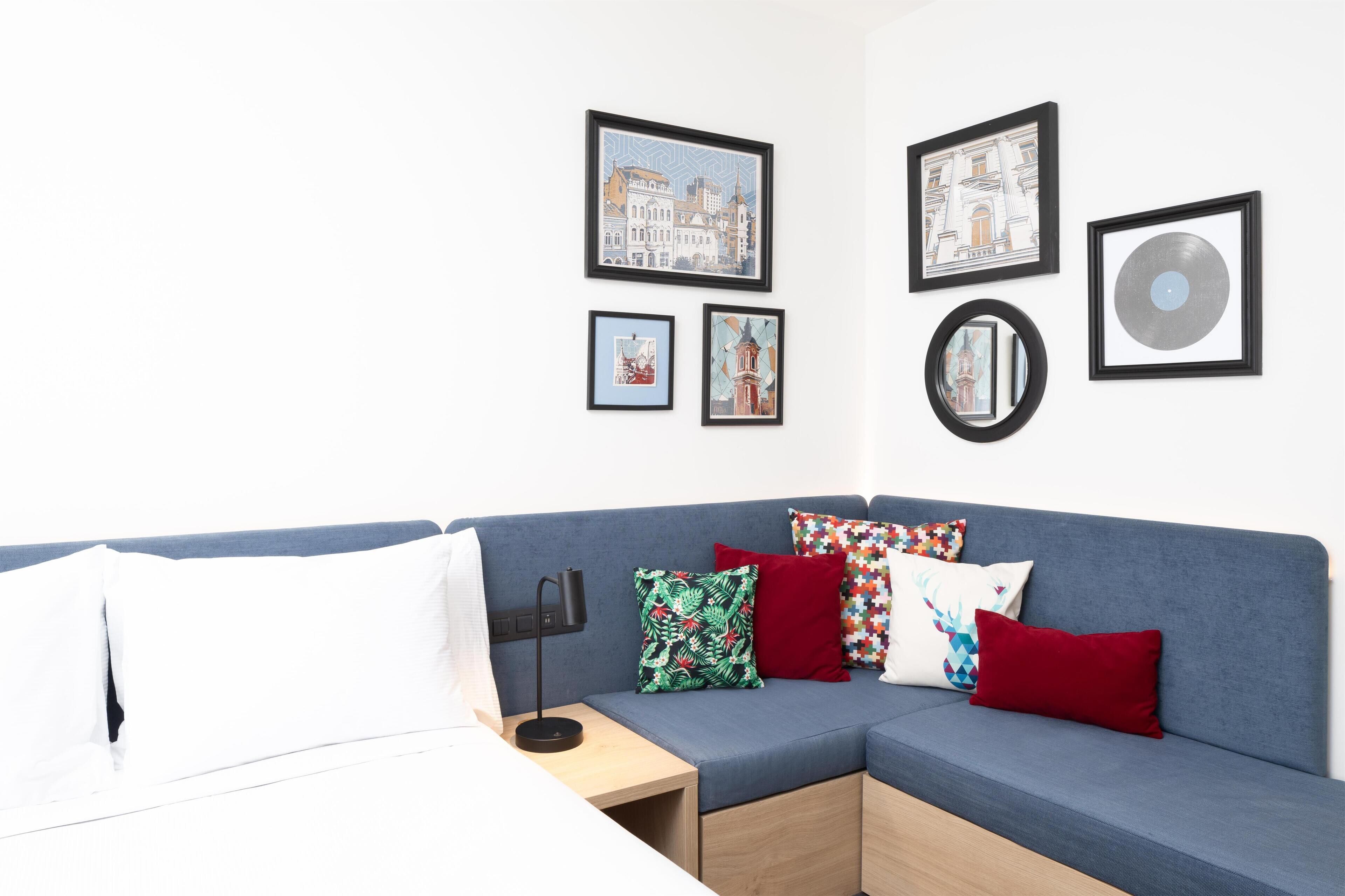 A modern room with a blue sofa, white pillows, and a small table with a lamp. There are framed pictures on the wall.