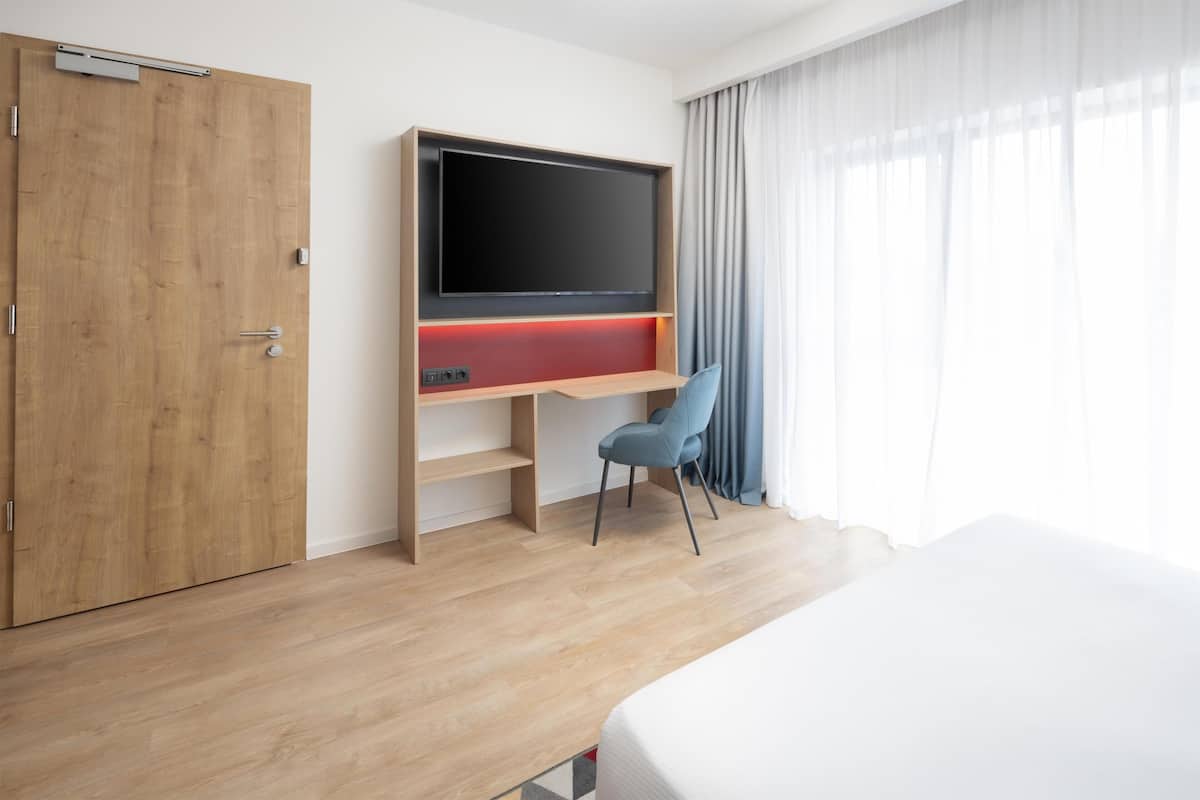 A hotel room with a wooden wardrobe, a TV mounted on a wooden shelf, a blue chair, and a bed.