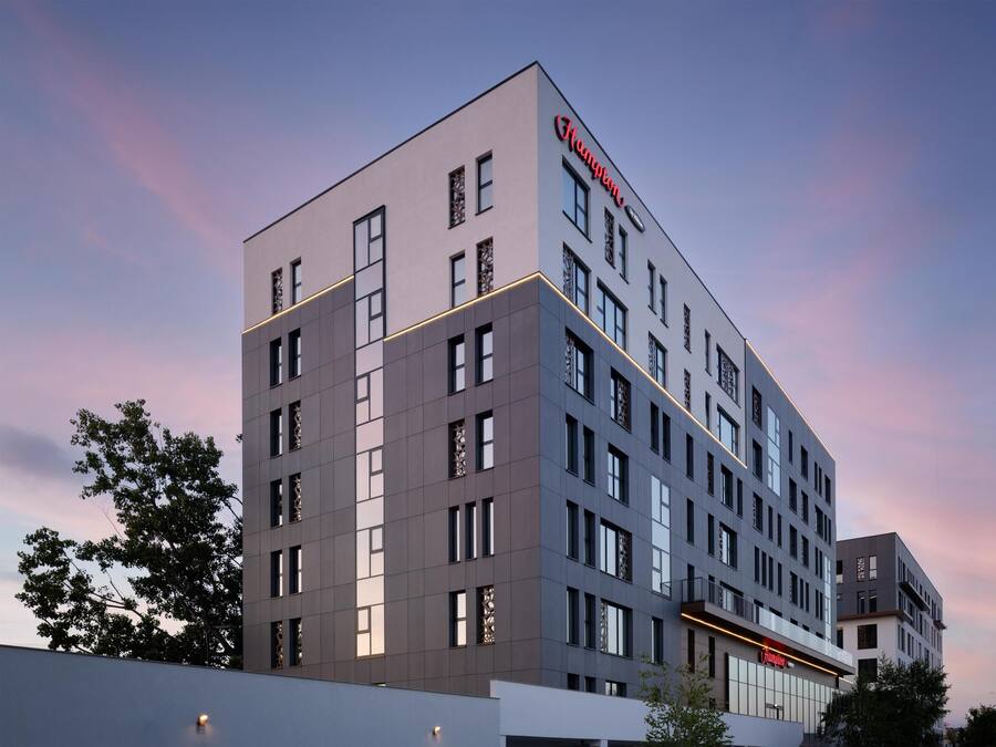 A modern hotel building with a prominent 'Hampton' sign, multiple floors, and a flat roof design.